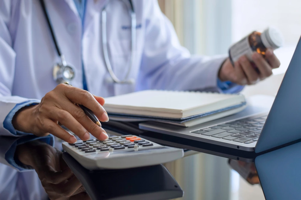 Doctor using a calculator at a desk with a pill bottle and laptop