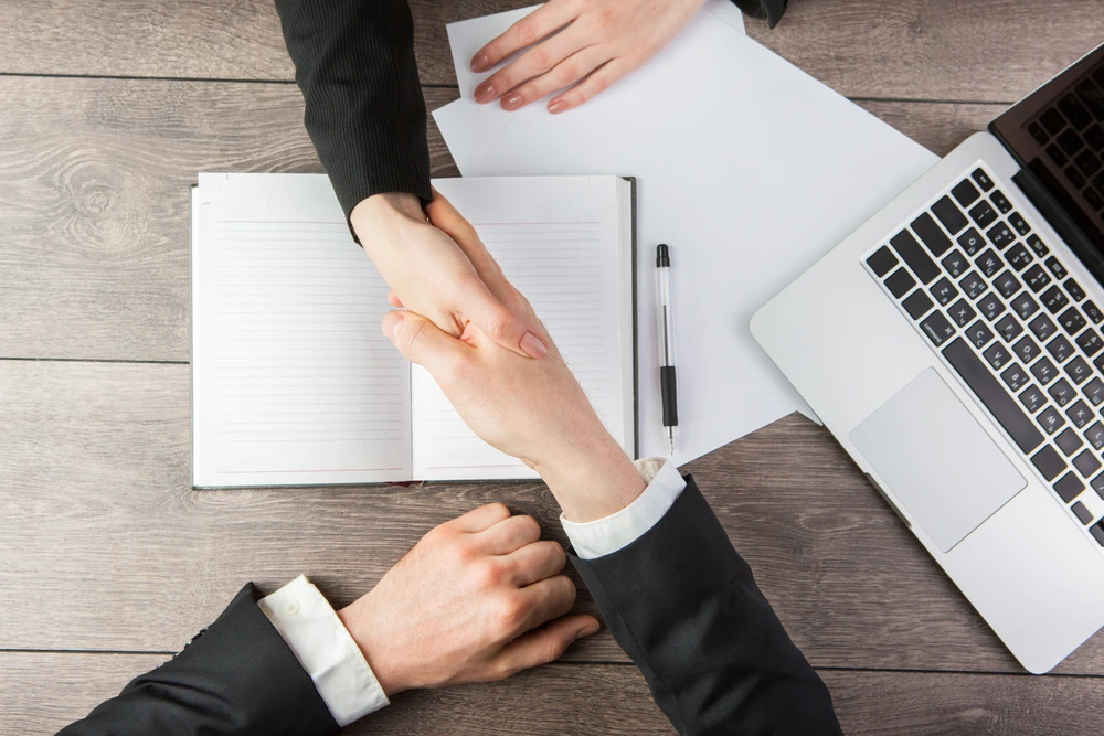 Handshake over a desk with a laptop and notebook